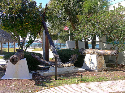 [Two rusted cannons sitting on white support structures on either side of a rusted anchor propped at an angle.]